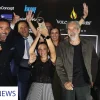 A group of six people, Imperio team, celebrates winning an award at the Cyprus Architecture, Interior Design & Property Awards 2024. A woman, Natalie, in the center raises the trophy high, surrounded by smiling and cheering teammates. The background features a black backdrop with sponsor logos, including "BoConcept," "Knauf," and "Volcano Stores."