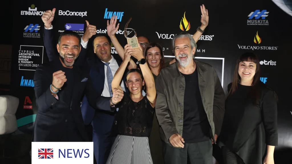 A group of six people, Imperio team, celebrates winning an award at the Cyprus Architecture, Interior Design & Property Awards 2024. A woman, Natalie, in the center raises the trophy high, surrounded by smiling and cheering teammates. The background features a black backdrop with sponsor logos, including "BoConcept," "Knauf," and "Volcano Stores."