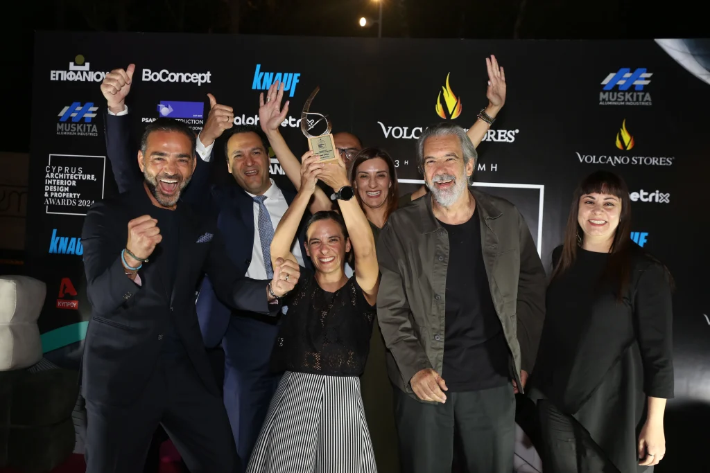 A group of six people, Imperio team, celebrates winning an award at the Cyprus Architecture, Interior Design & Property Awards 2024. A woman, Natalie, in the center raises the trophy high, surrounded by smiling and cheering teammates. The background features a black backdrop with sponsor logos, including "BoConcept," "Knauf," and "Volcano Stores."